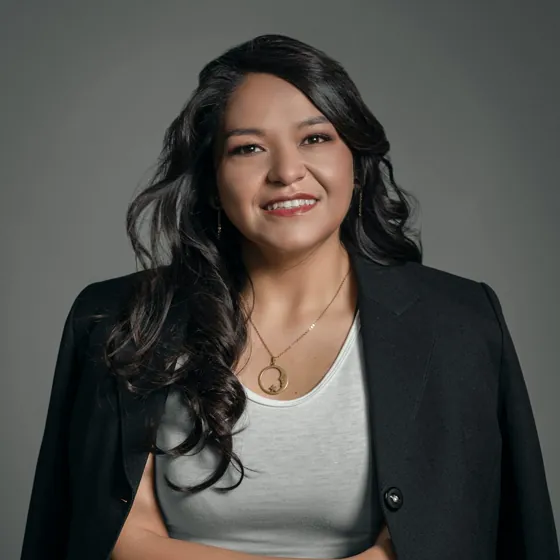 A smiling woman with long dark hair in a black blazer and white top, against a grey backdrop.