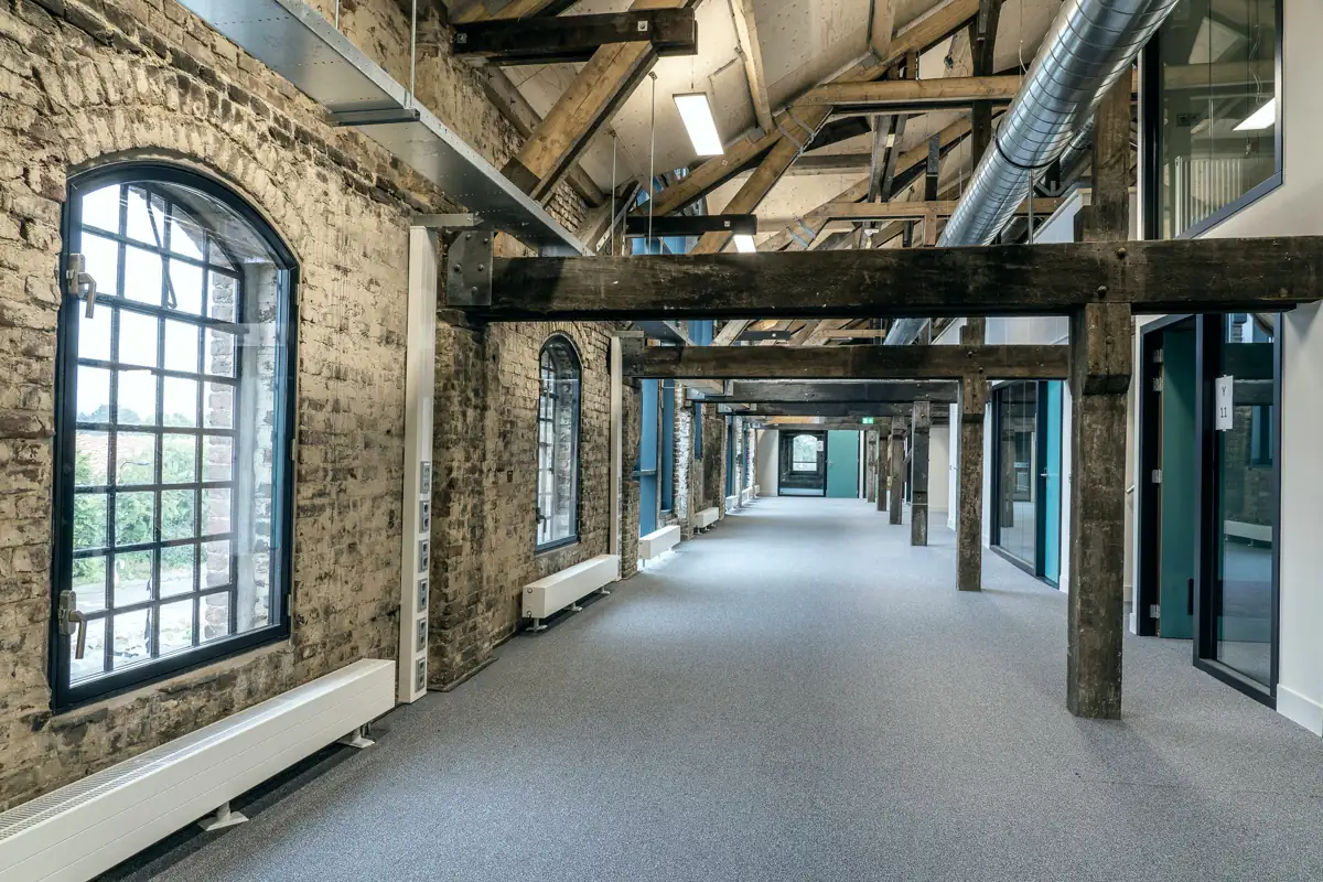 A wide corridor with arched steel windows in the original brick wall, timber beams overhead and glass office fronts on the right.
