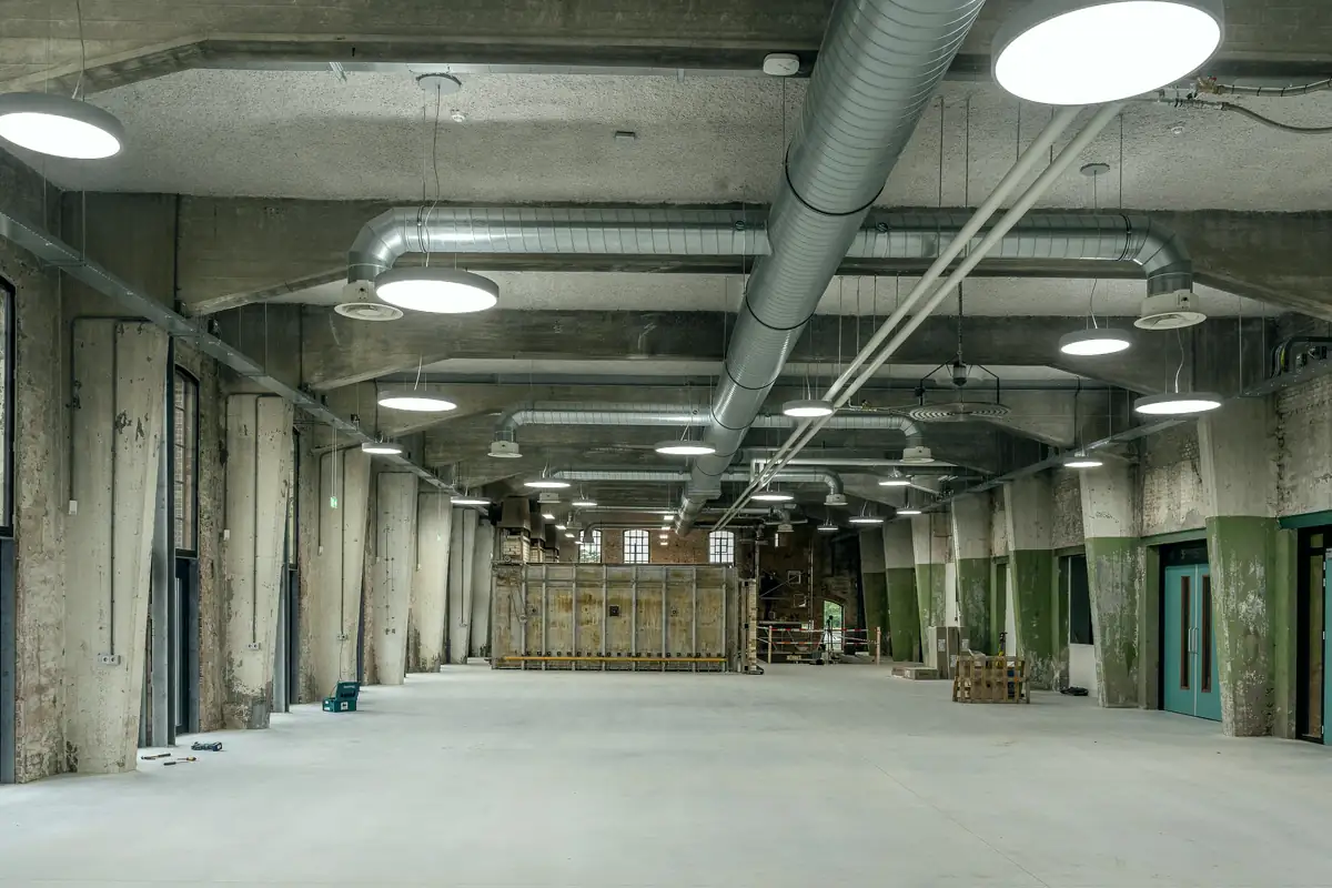 An open ground-floor space with a concrete floor, exposed ducts, round ceiling lights and tall windows along one side.