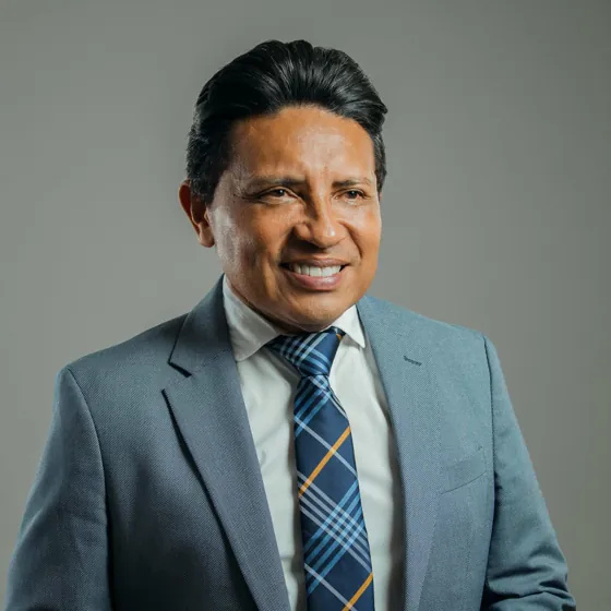 A smiling man in a light blue suit and plaid tie, against a grey backdrop.