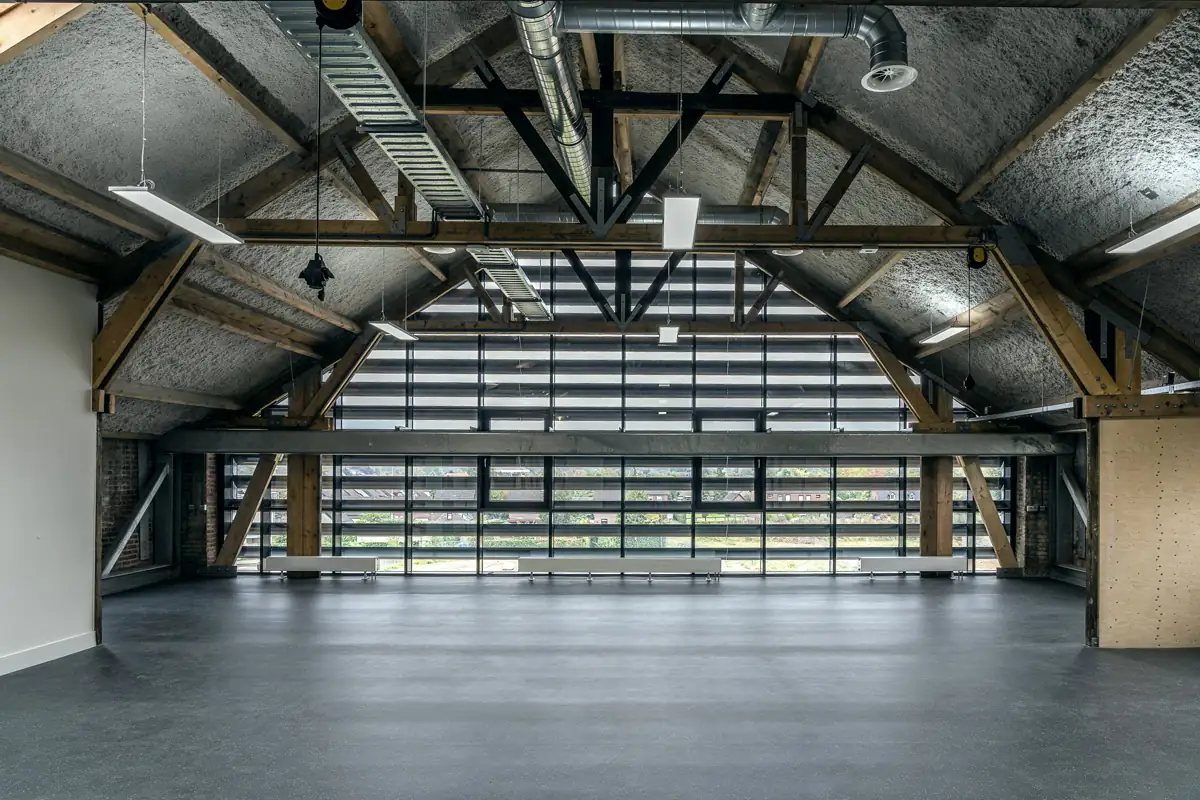 An empty upper floor under exposed timber roof trusses, with a polished concrete floor and a full-width window wall at the far end.