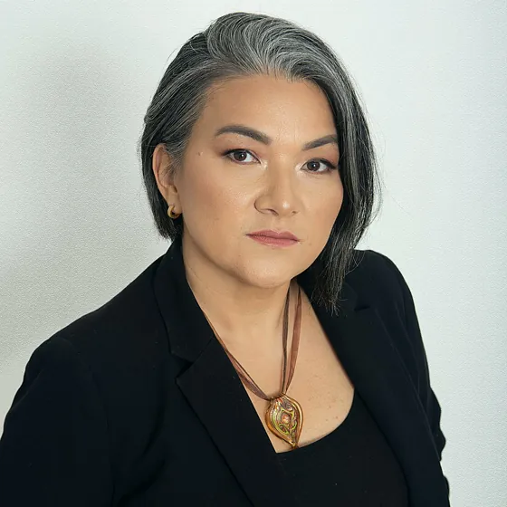 A woman with short grey hair in a black jacket and a pendant necklace, against a pale grey backdrop.