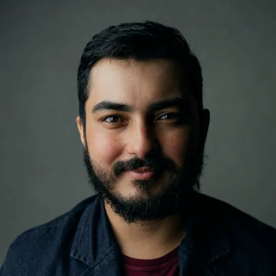 A man with a dark beard in a navy blazer over a deep red shirt, against a grey backdrop.